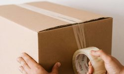 Crop anonymous male using tape to seal packed cardboard box against white wall while moving into new place