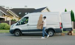 Man unloading cardboard boxes from a delivery van in a suburban neighborhood street.